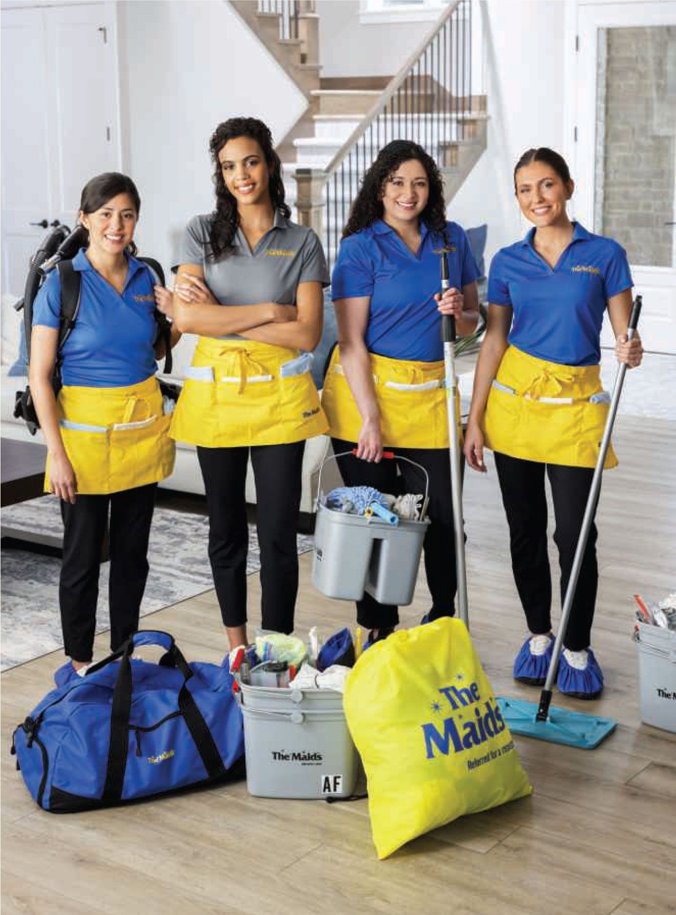 Four cleaning staff in blue uniforms with yellow aprons stand with mops and buckets in a bright hallway.