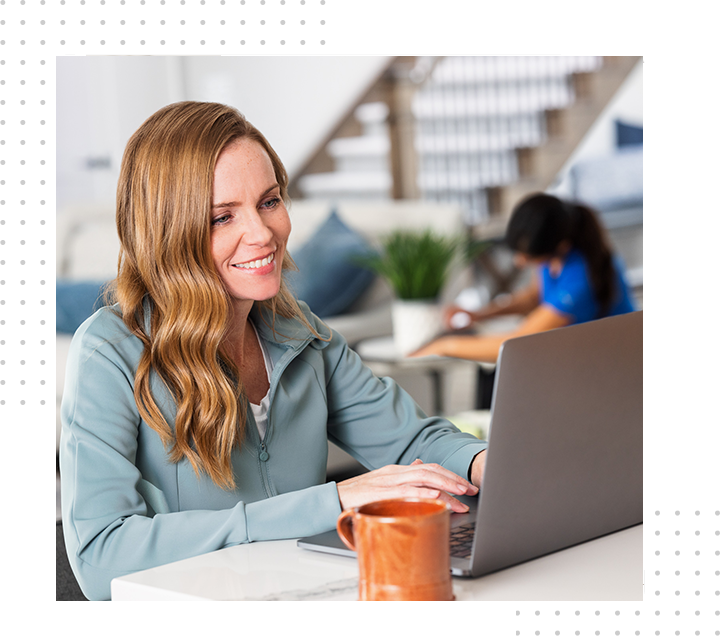 Smiling woman using a laptop at a café table, with an orange mug in front and a blurred coworker behind her