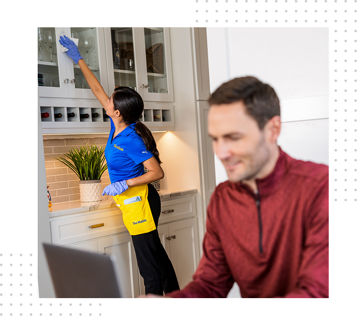 Cleaner reaching for a cabinet in a bright kitchen while a man works in the foreground