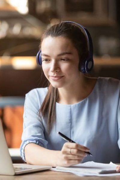 Woman wearing headphones, focused, taking notes while looking at a laptop in a cafe.
