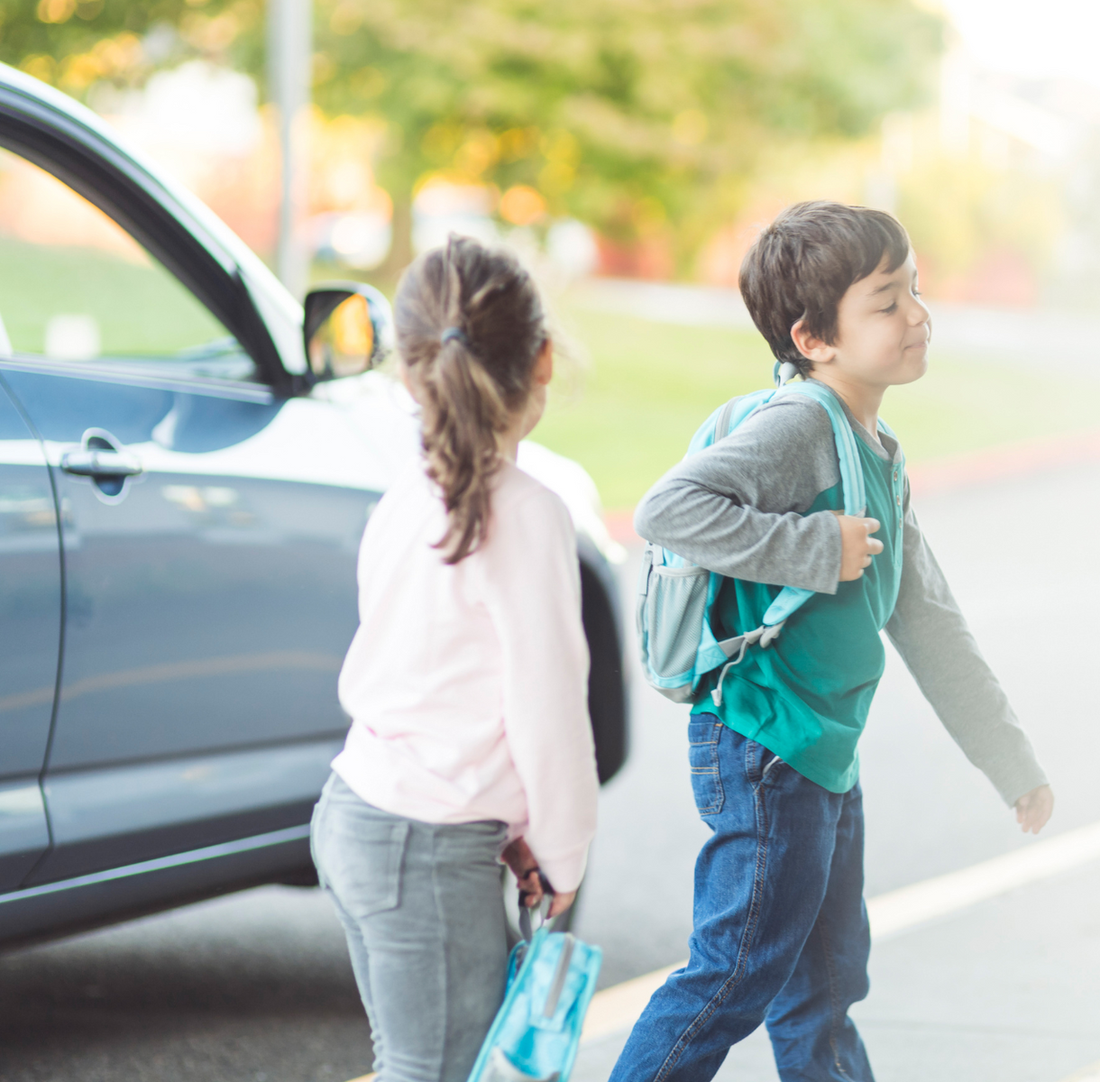 Two children with backpacks walking away beside an open car in a bright driveway