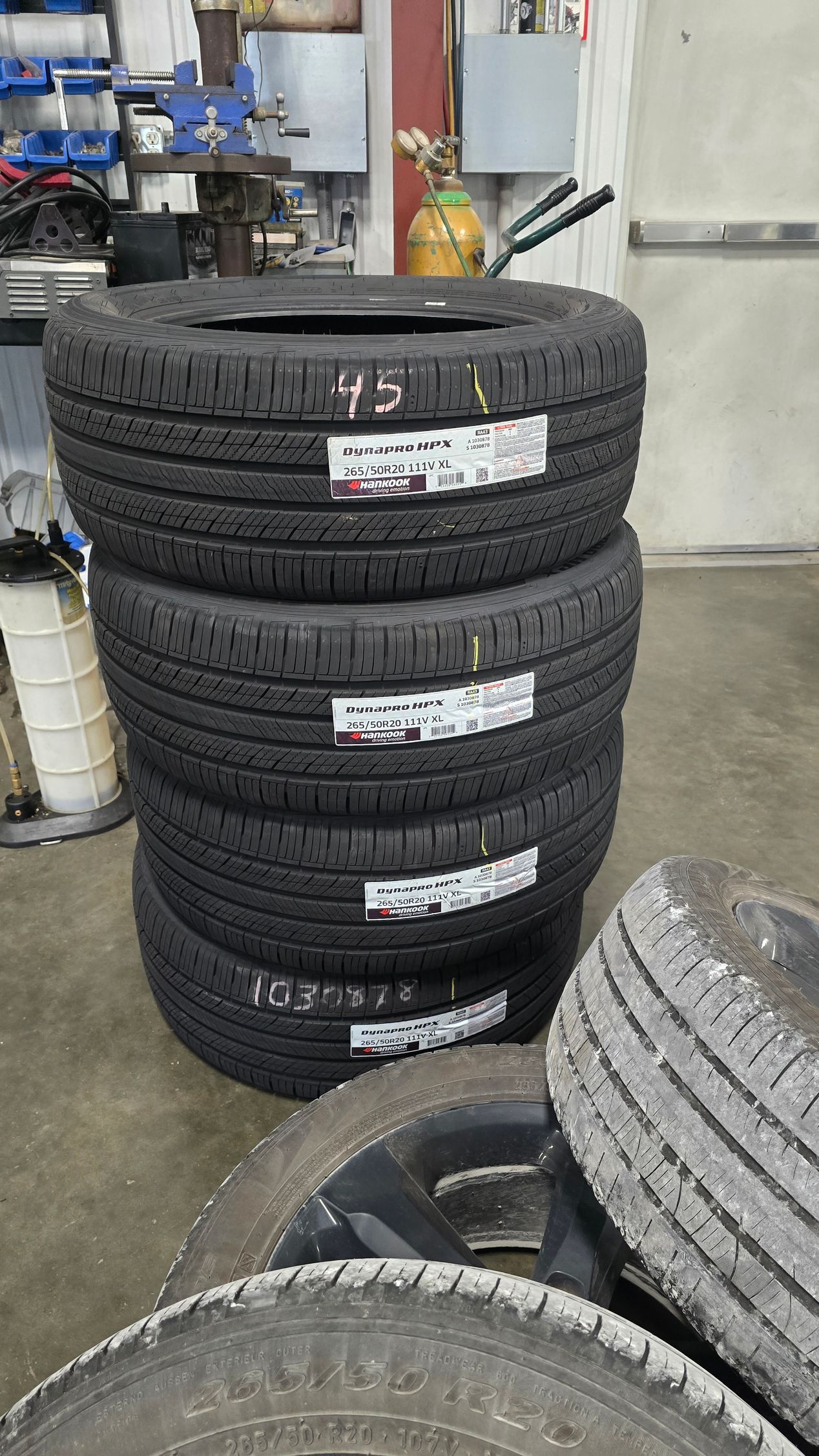 Stacks of black car tires in a workshop, with a partial tire stack in the foreground and garage equipment behind them.