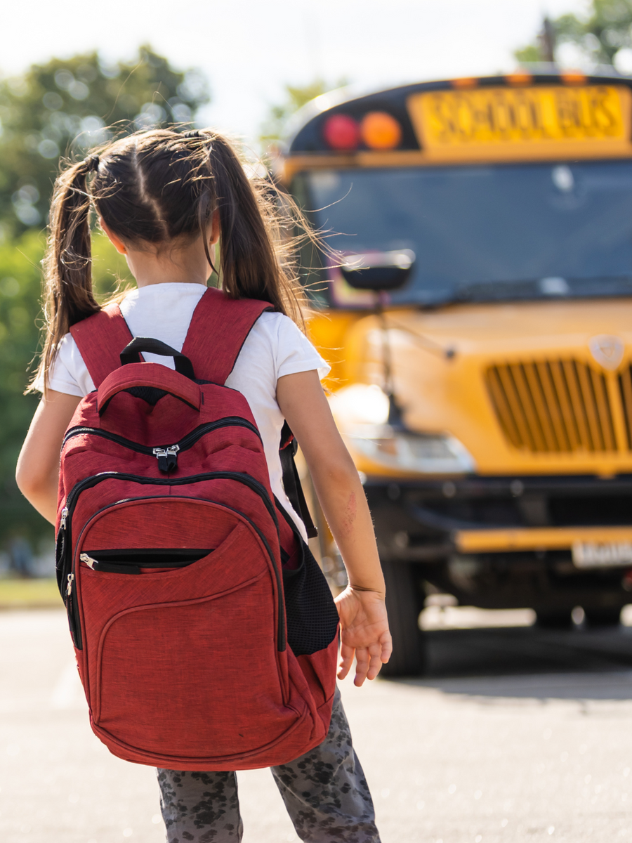 Child with red backpack walking toward a yellow school bus on a street