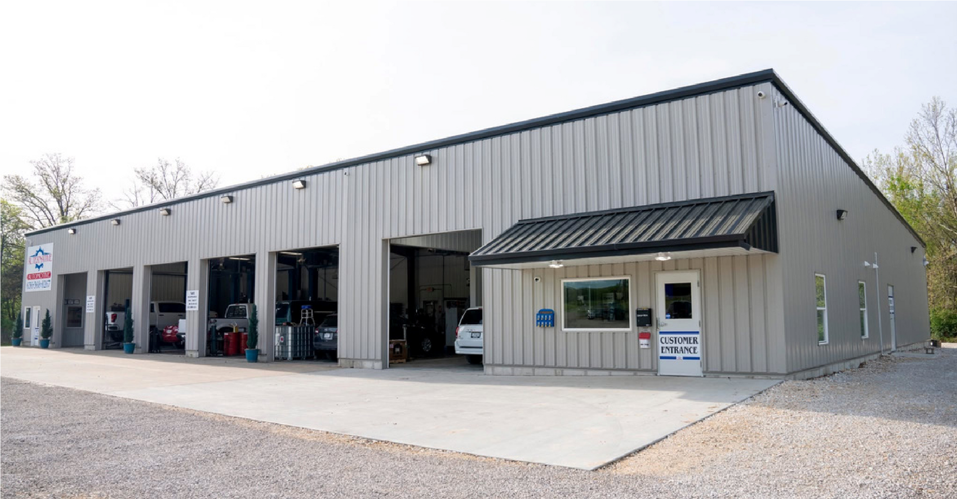 Gray metal garage building with open service bays and a small office entrance on a gravel lot