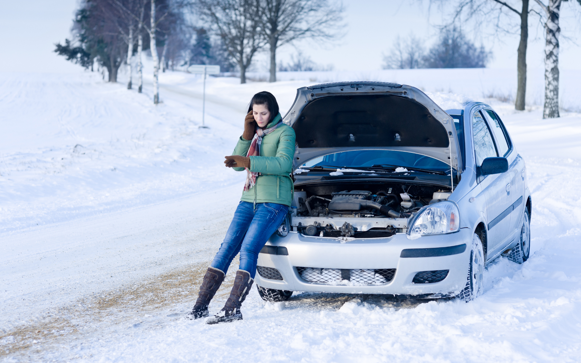 Woman leaning against a silver car with open hood on a snowy road, checking her phone