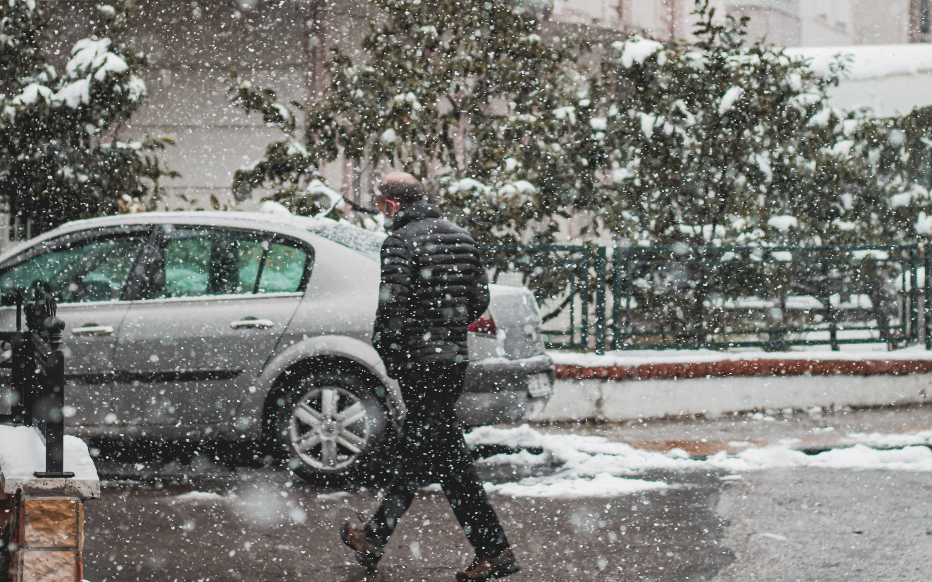 Person walking in heavy snow past a parked silver car and trees.