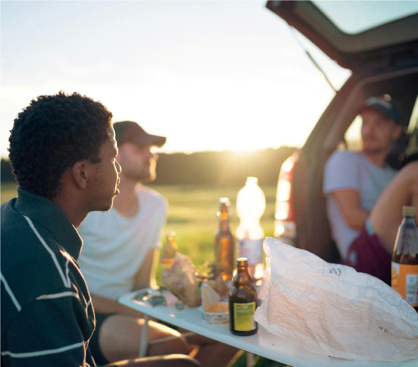 Friends chatting beside an open car trunk at sunset with drinks and snacks on a picnic table.
