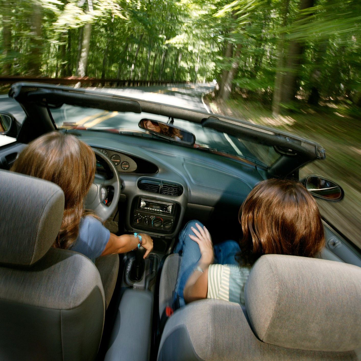 Two people riding in a convertible on a winding forest road, viewed from the back seat