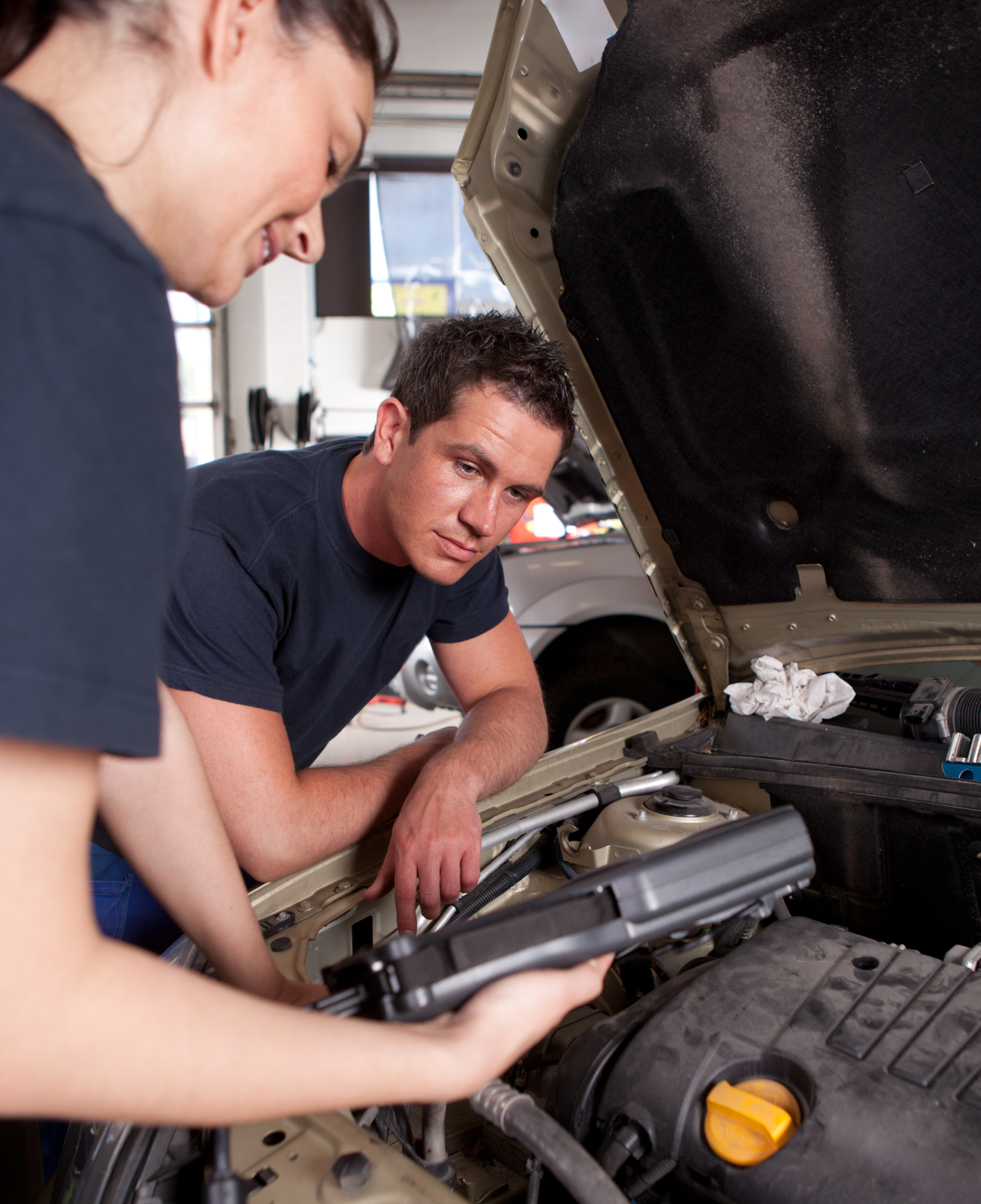 Two mechanics inspecting an open car engine in a garage, one holding tools near the engine bay.