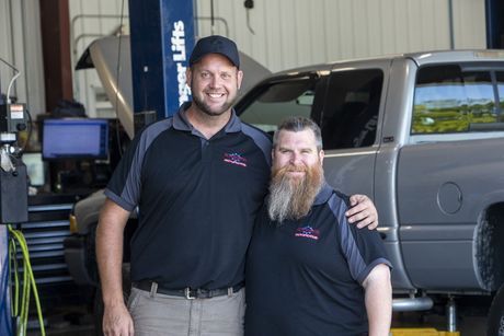 Two mechanics smiling beside a silver SUV in an auto repair shop
