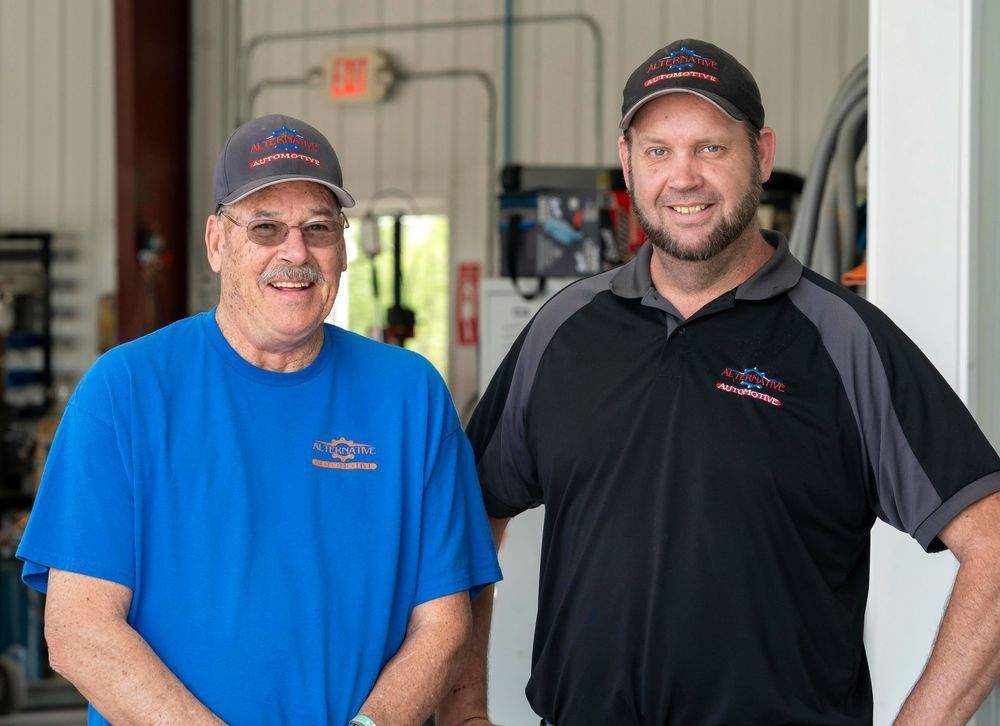 Two smiling employees standing in a garage, one in a blue shirt and one in a black shirt.