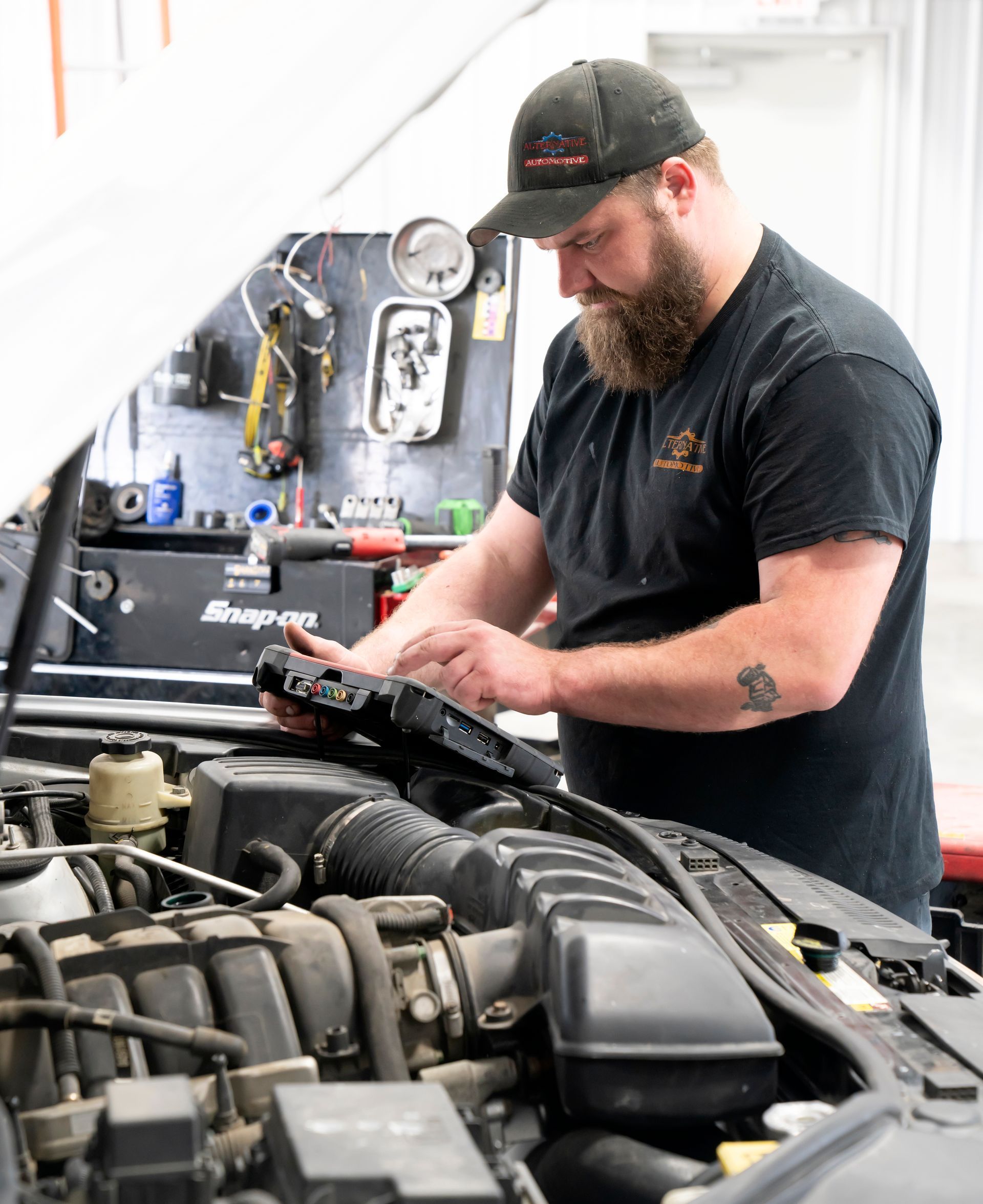 Mechanic inspecting a car engine in a workshop, wearing a black cap and shirt