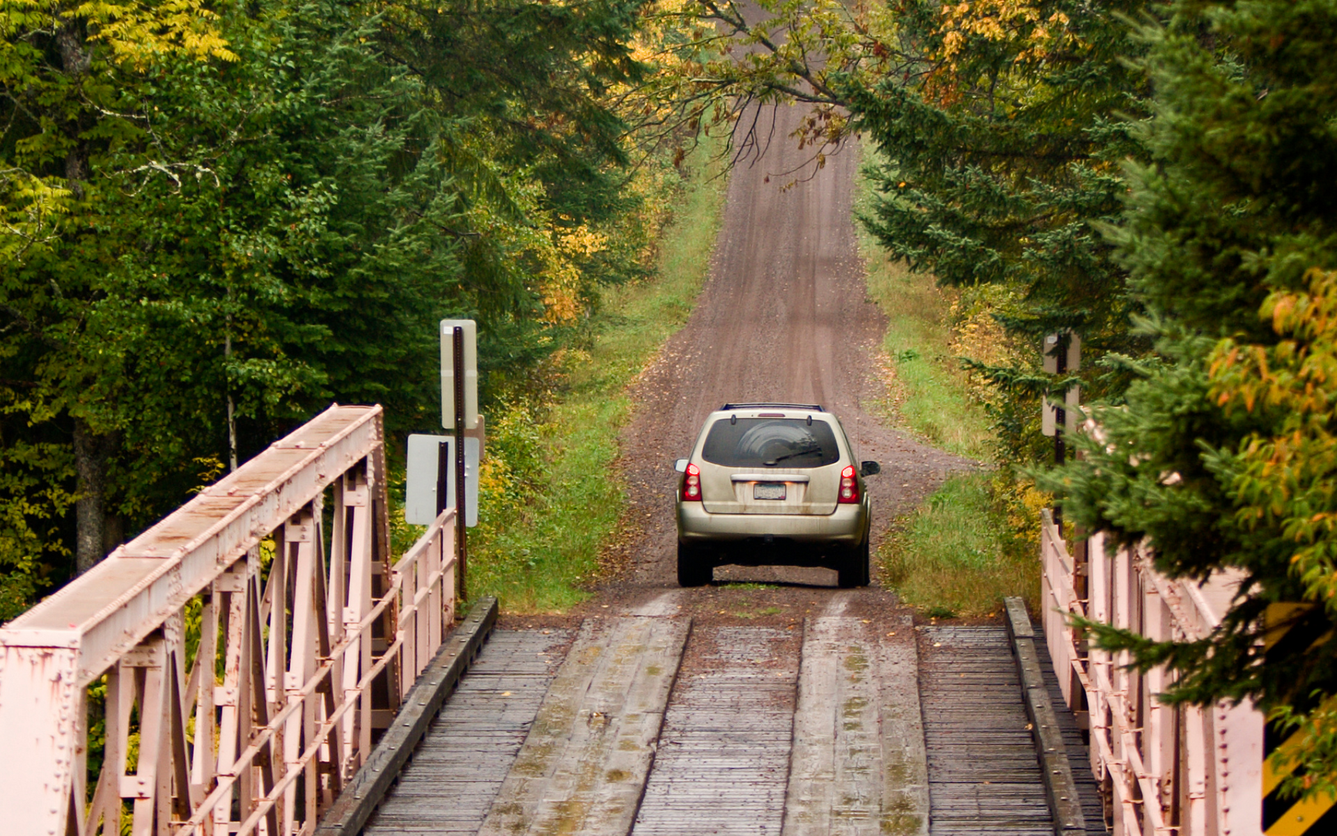 White SUV on a narrow wooden bridge leading to a dirt road through green trees.