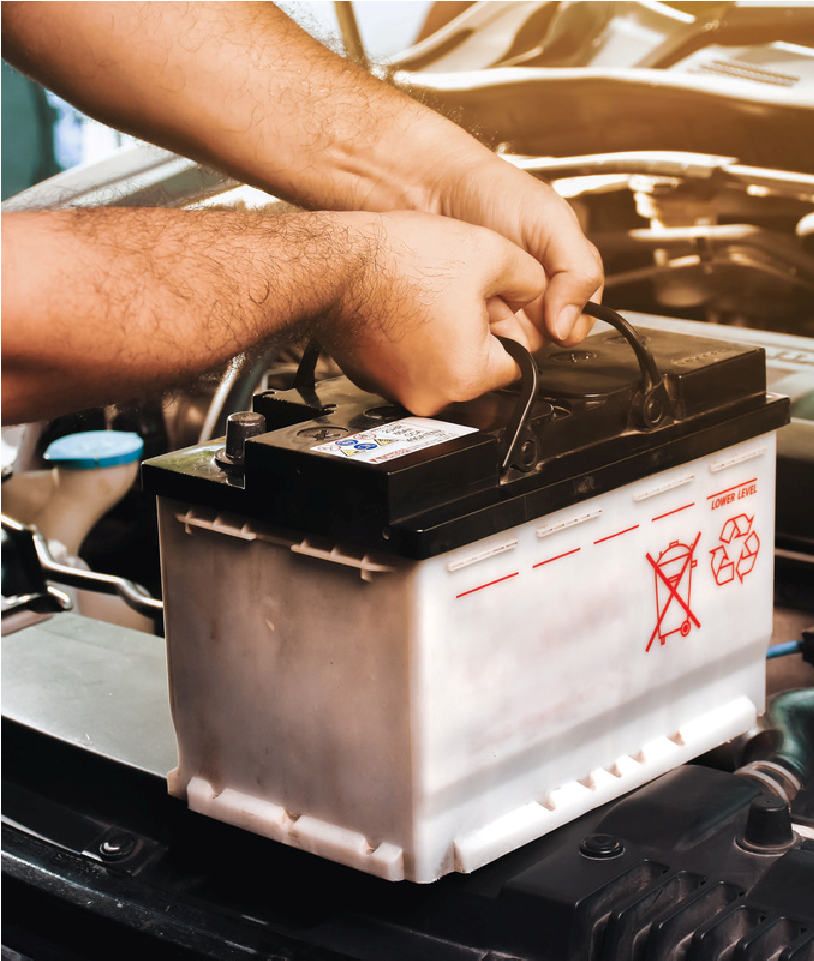 Hands lifting a car battery in an engine bay, with red warning symbols on the side.