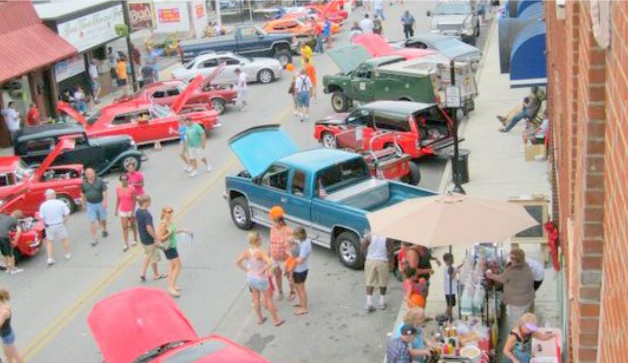 Classic car show on a crowded street, with red, blue, and green vintage vehicles and pedestrians browsing.