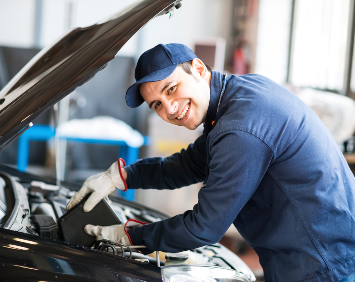 Mechanic in blue uniform smiling while inspecting a car engine with the hood open