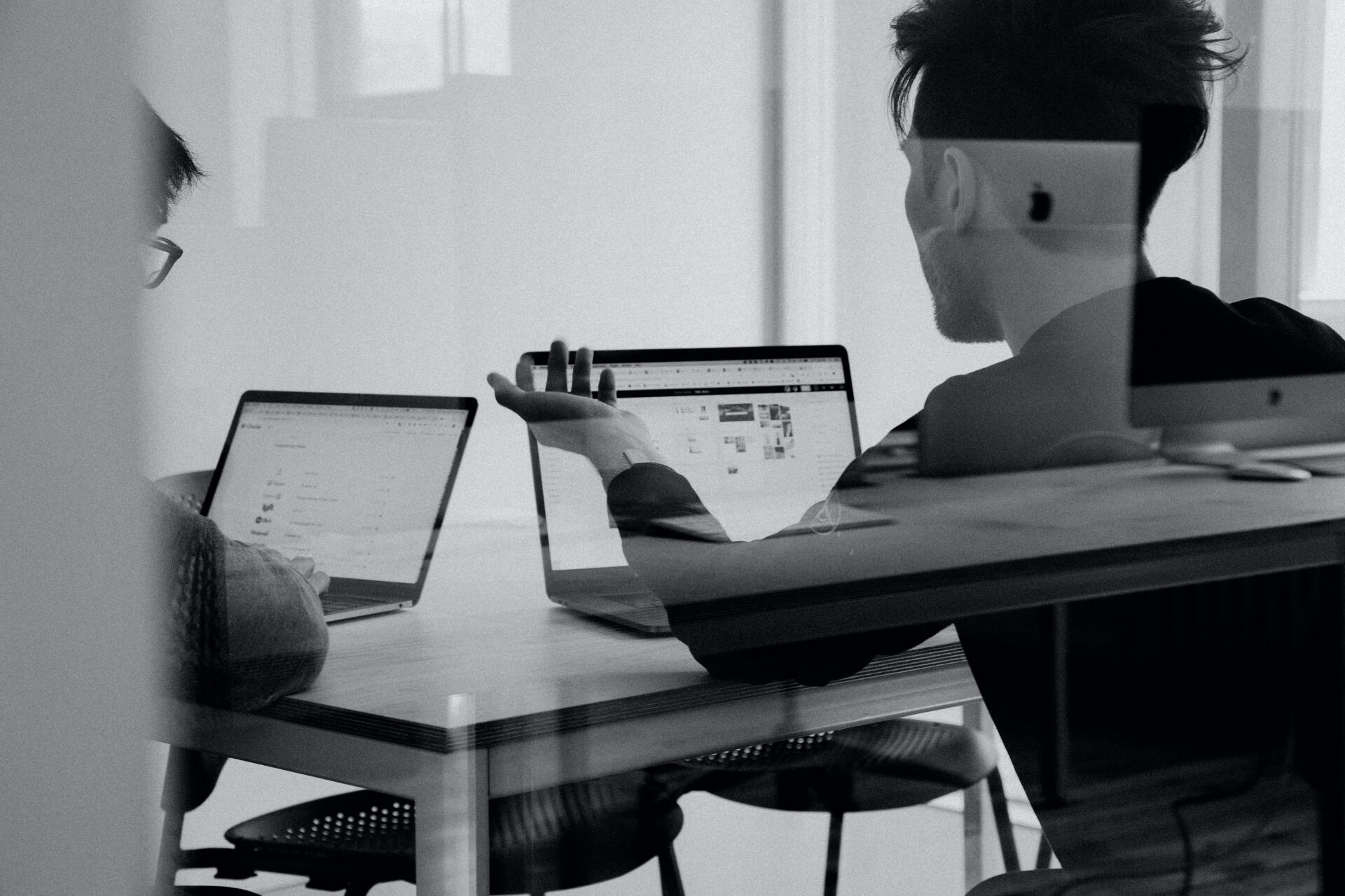 A black and white photo of two people sitting at a table with laptops.