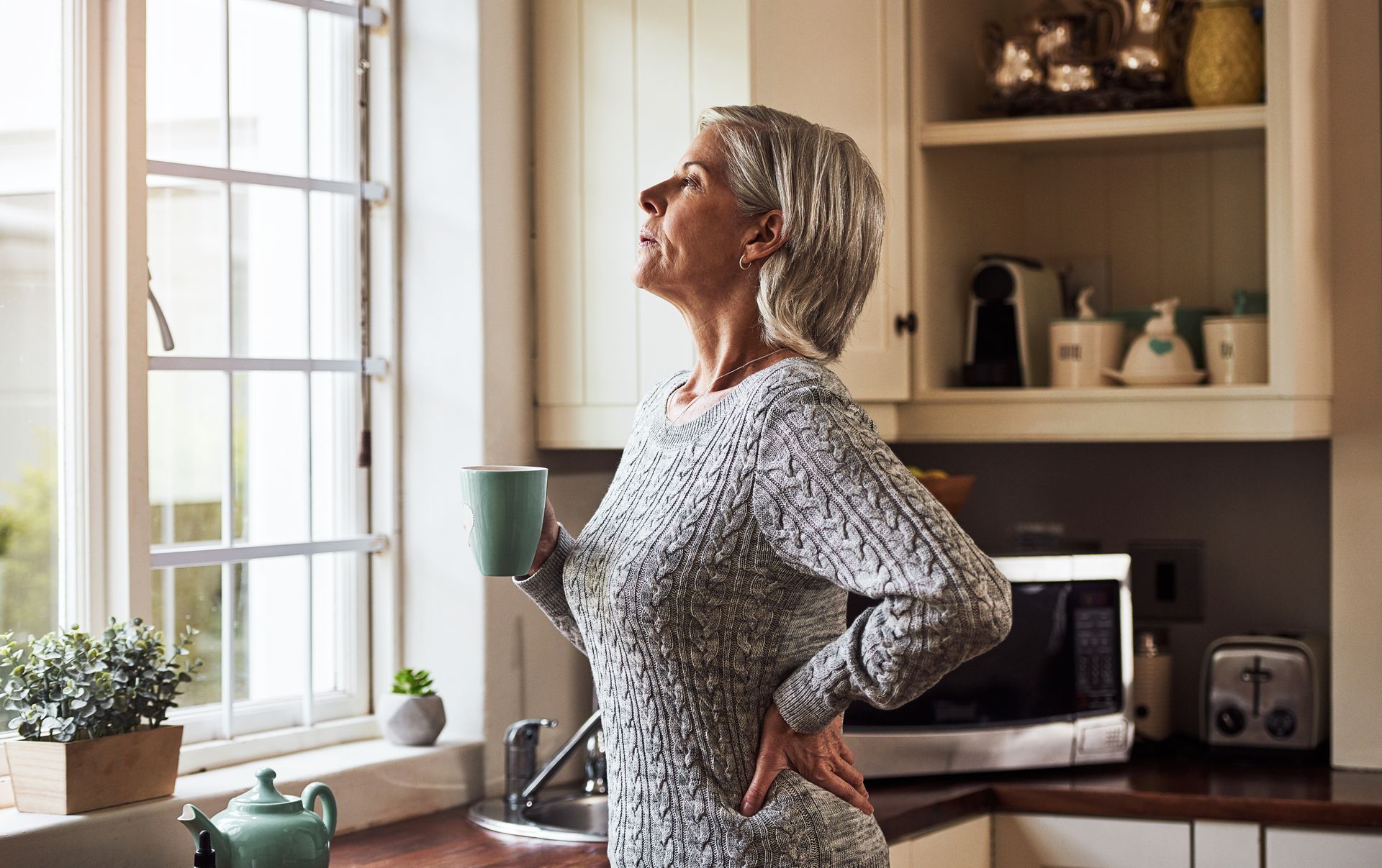 Woman stretching at a kitchen window, holding a mug in a bright, modern kitchen.