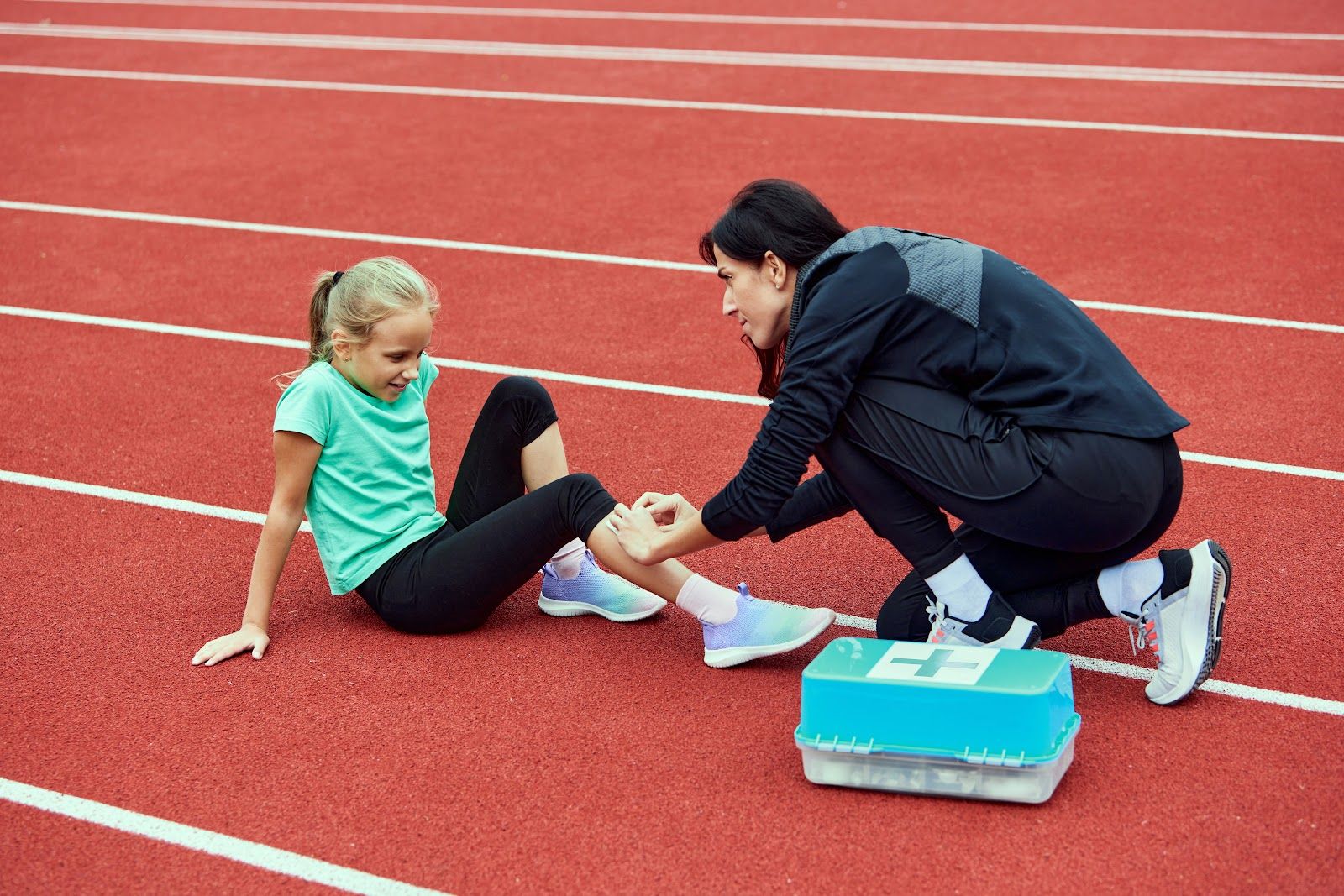 Coach tending to an athlete's ankle on a red track beside a blue first-aid kit