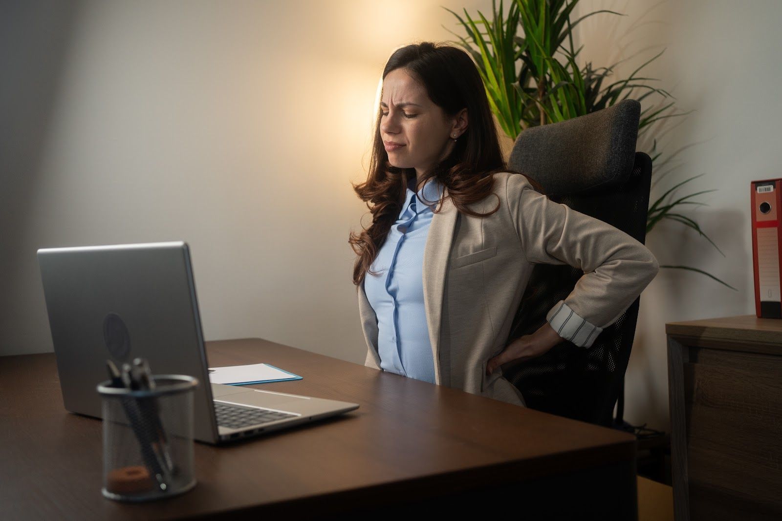 Woman sitting at a desk, looking at a laptop, with a plant and lamp in the background.
