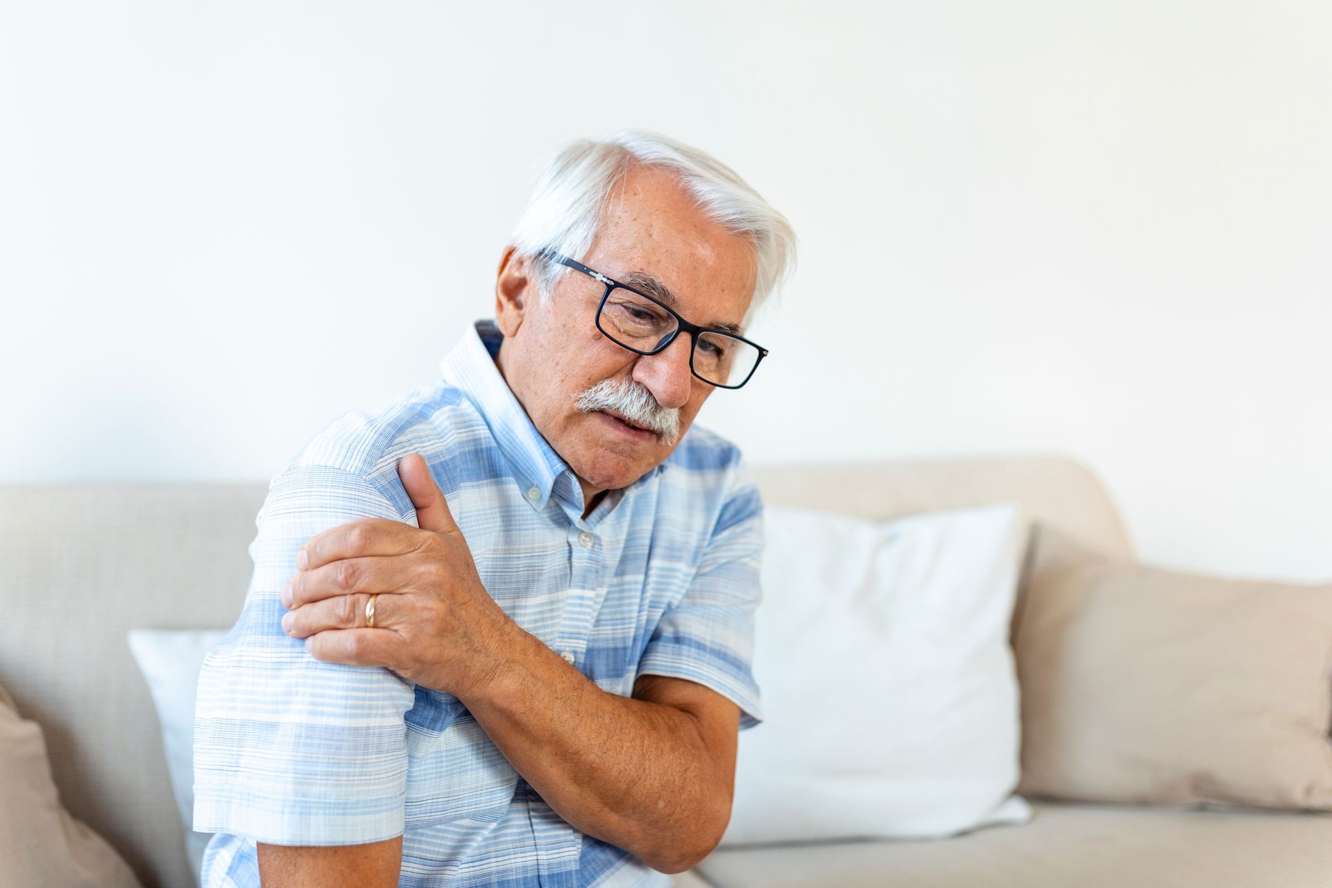 Elderly man in glasses holding his shoulder while sitting on a couch, looking uncomfortable.