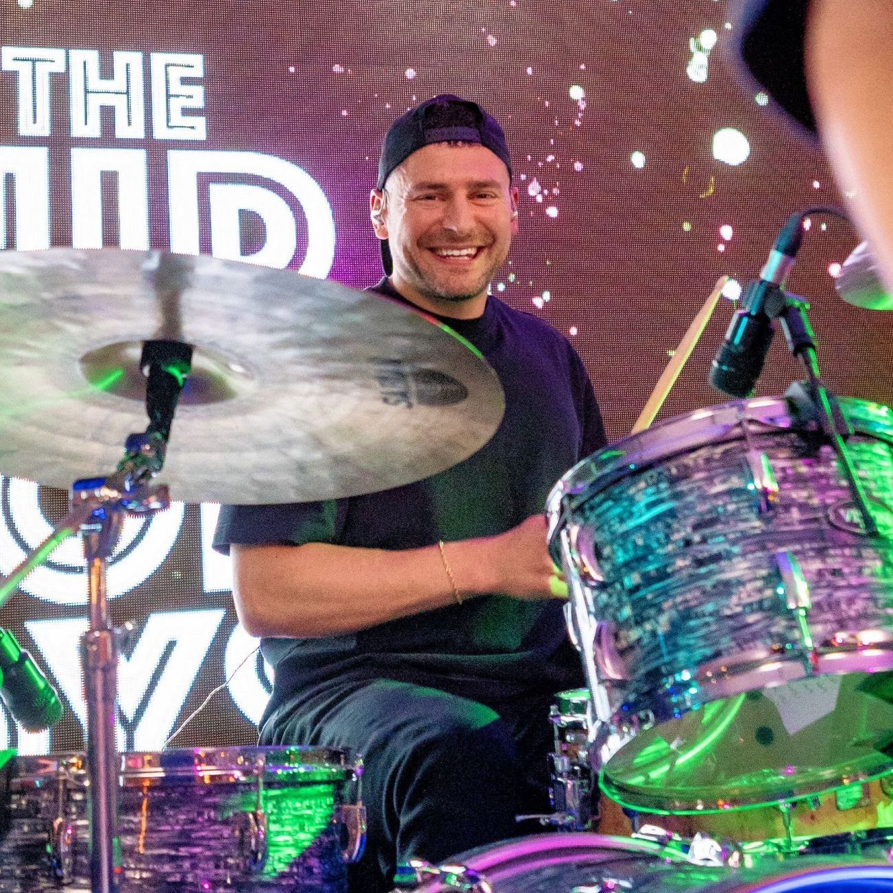 Drummer in a black cap and shirt, smiling while playing a turquoise drum set. Stage setting.