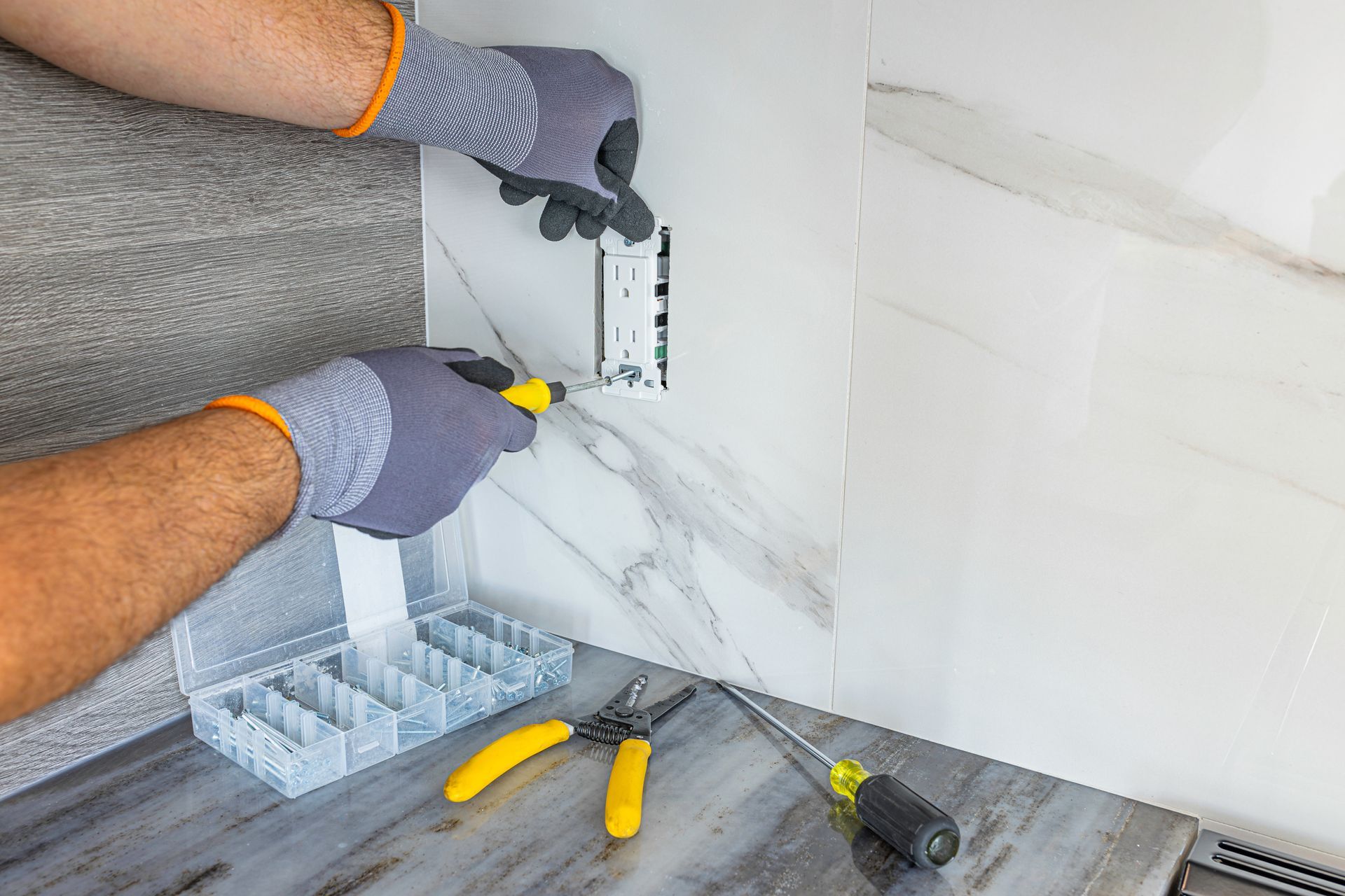 Hands installing a wall outlet in a kitchen with tools and tile backsplash