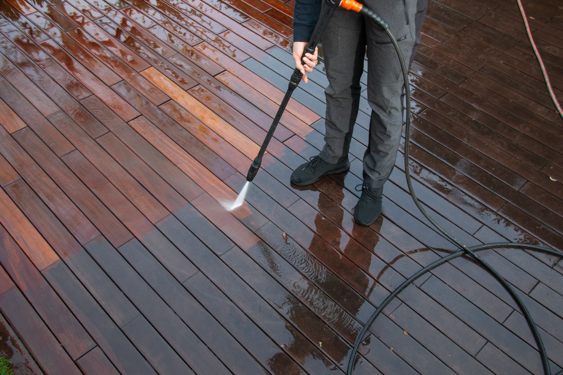 Person pressure-washing a wet wooden deck with a hose nozzle and long wand