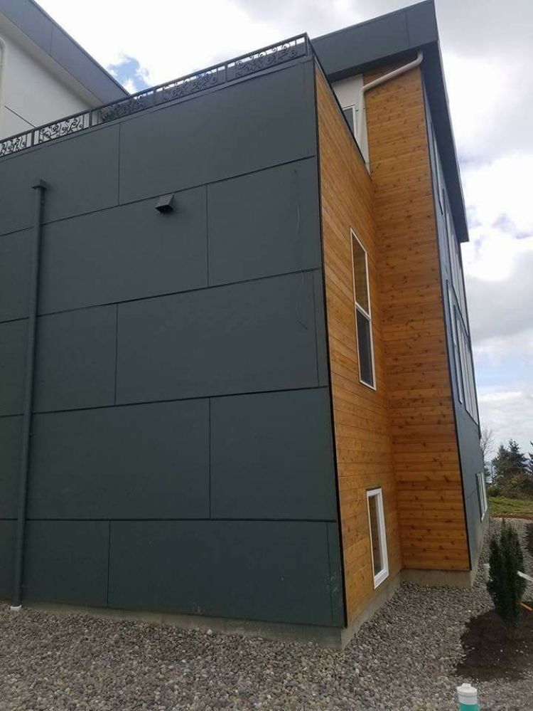 A modern building exterior featuring dark gray paneling on one side and horizontal wood siding on the other, set in gravel.
