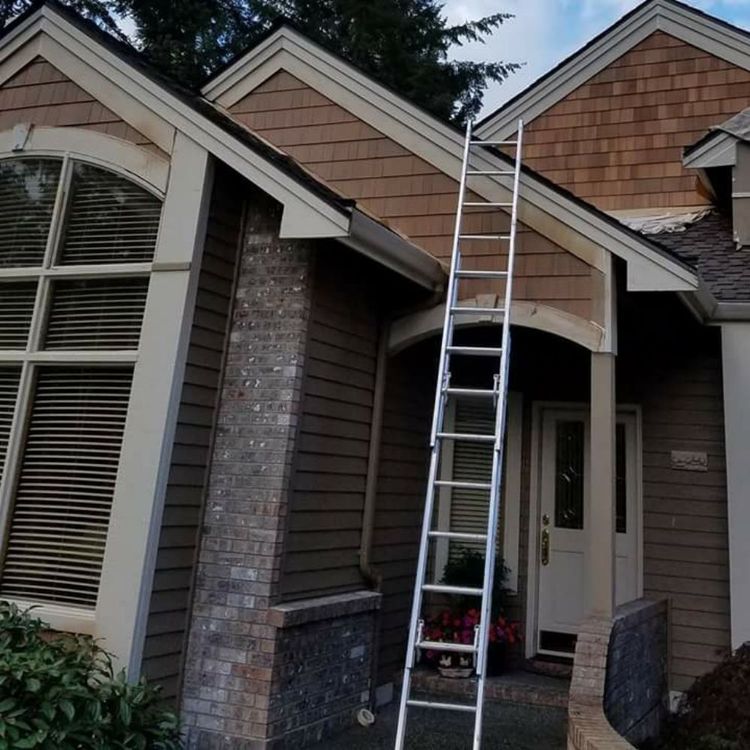 An aluminum extension ladder leans against the shingled roofline above the entrance of a house with stone and siding.