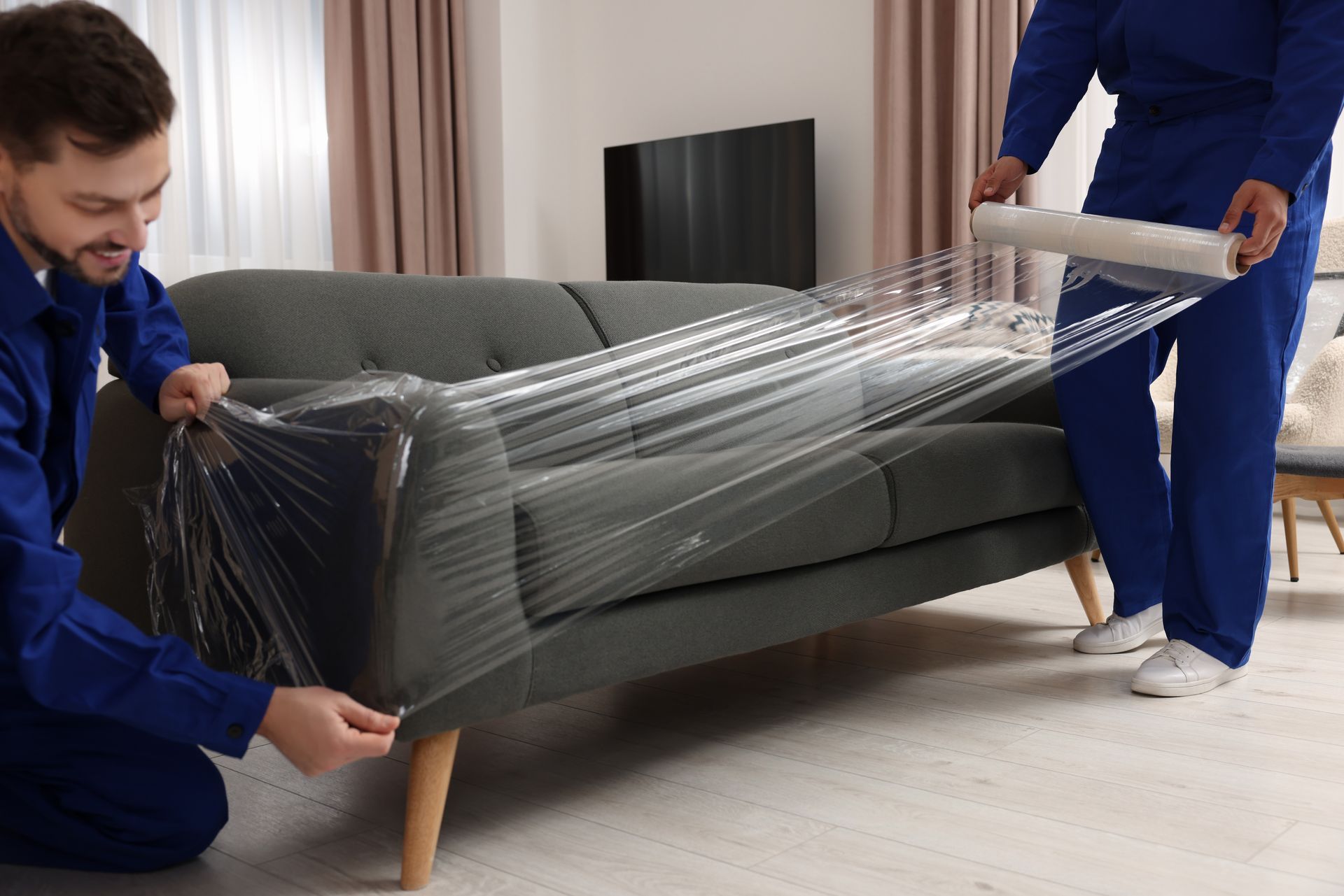 A male mover with stretch film wrapping a sofa in a living room with a co-worker.