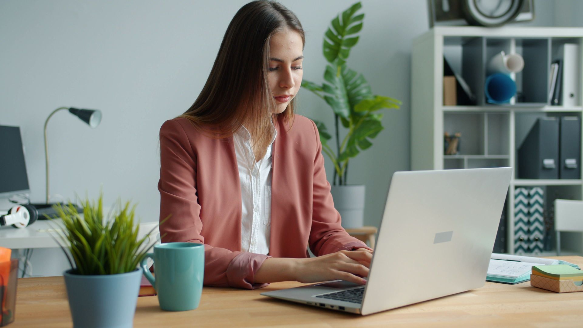 Una donna digita al computer portatile seduta a una scrivania in un ufficio luminoso, con una tazza, una pianta e degli scaffali nelle vicinanze.