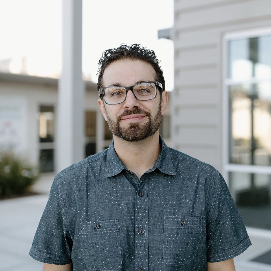 A man with a beard and glasses is standing in front of a building.