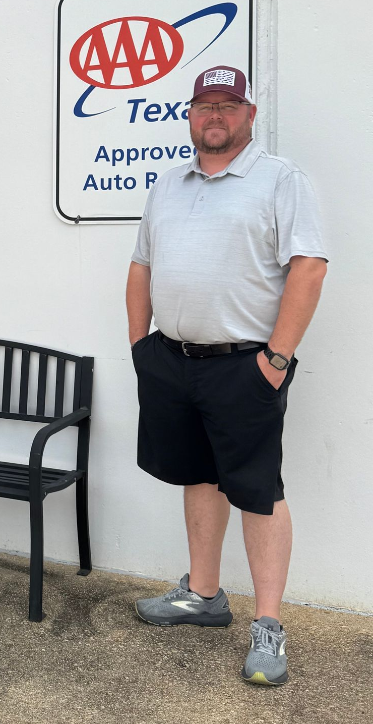 Man standing by a AAA sign, wearing a gray polo, black shorts, cap, and sunglasses, with hand in pocket.
