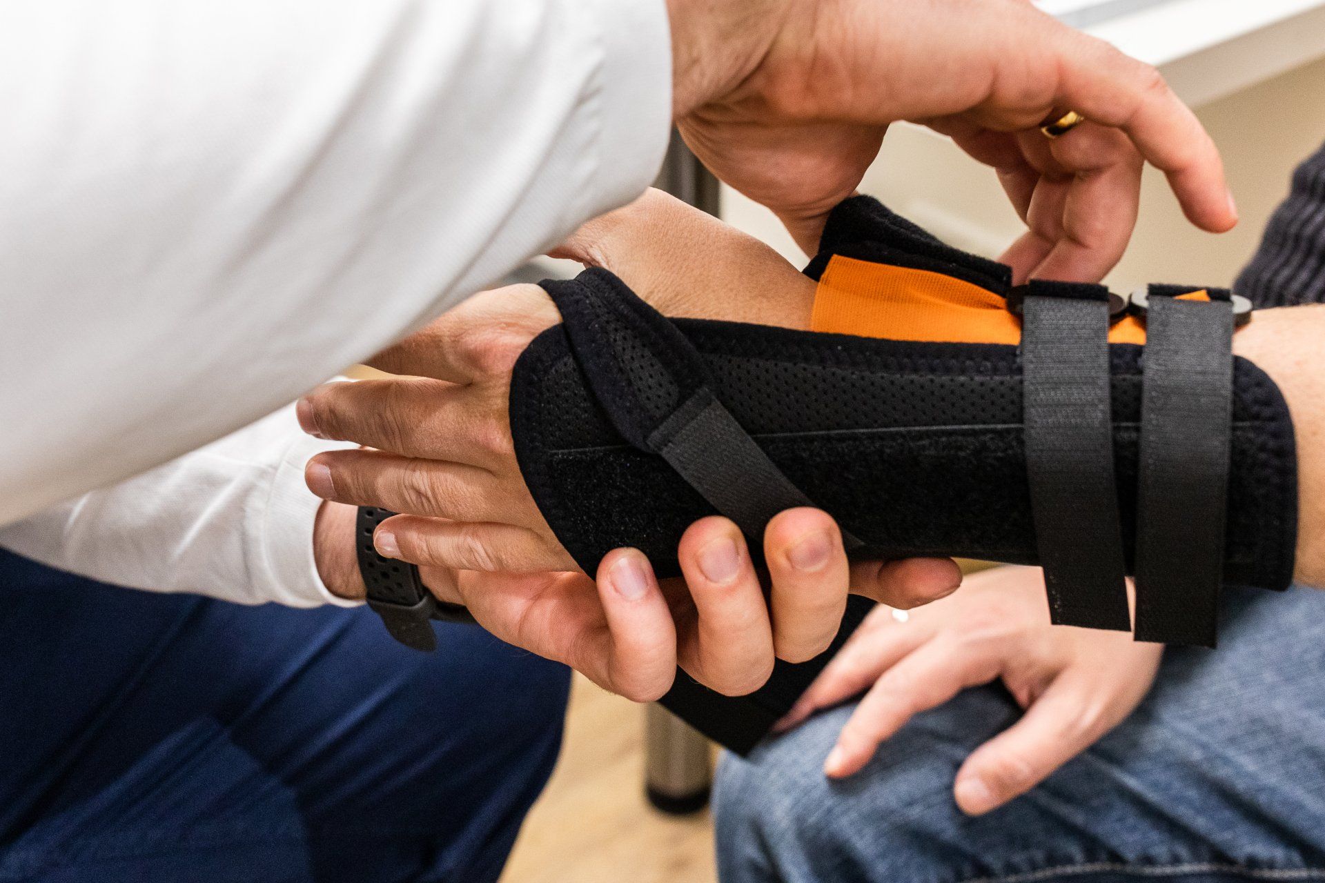 Ein medizinischer Fachmann befestigt eine schwarze Handgelenkschiene mit orangefarbenem Futter an der Hand eines Patienten.