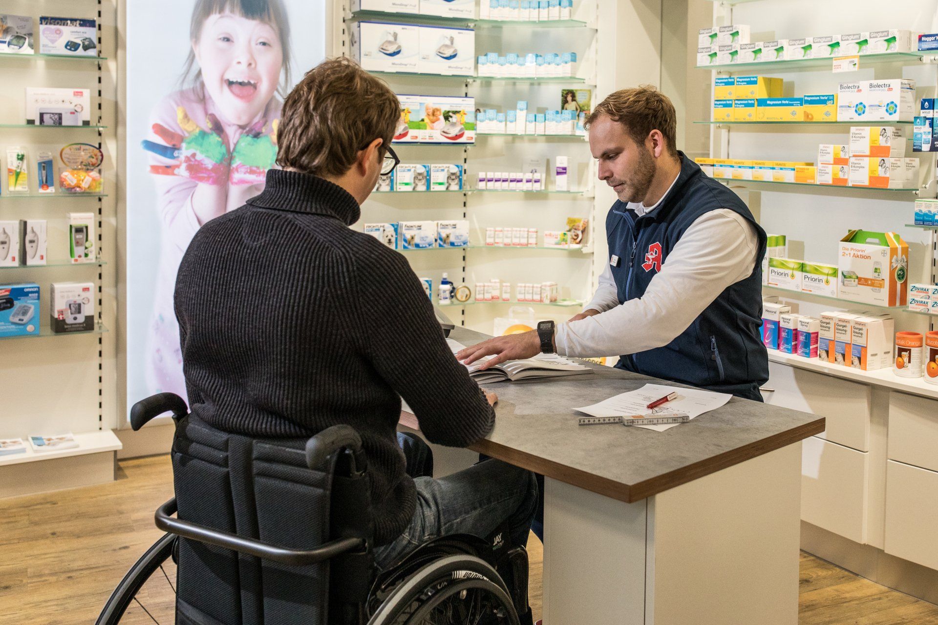 Eine Person im Rollstuhl berät sich mit einem Apotheker an einem Schalter in einer Apotheke, die mit Gesundheitsprodukten bestückt ist.