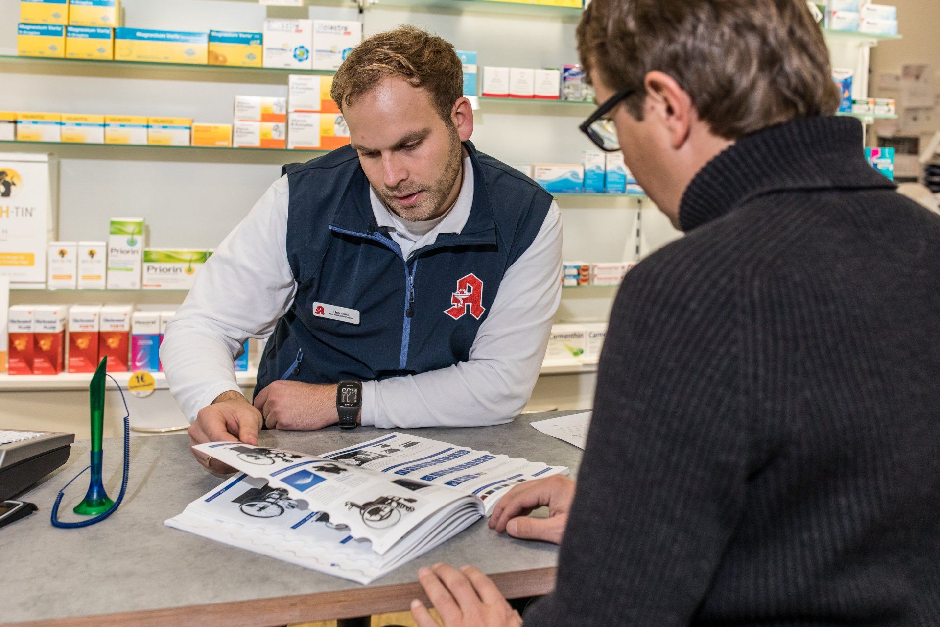 Ein Apotheker in blauer Weste bespricht ein Dokument mit einem Kunden an einem Apothekentresen, dessen Regale mit Medikamenten gefüllt sind.