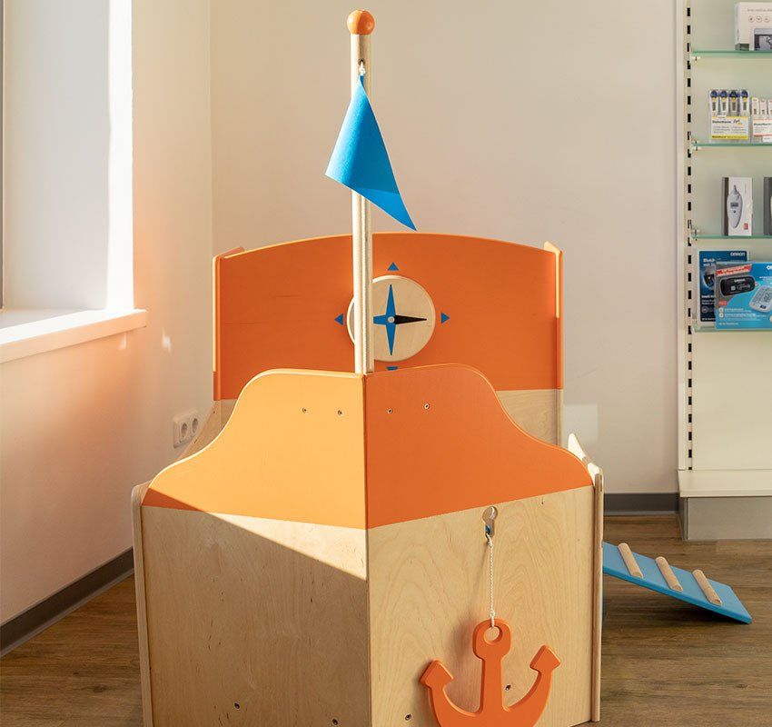 Ein hölzernes Kinderspielgerät in Form eines Bootes mit orangefarbenem Deck, Mast mit blauer Flagge und Anker.