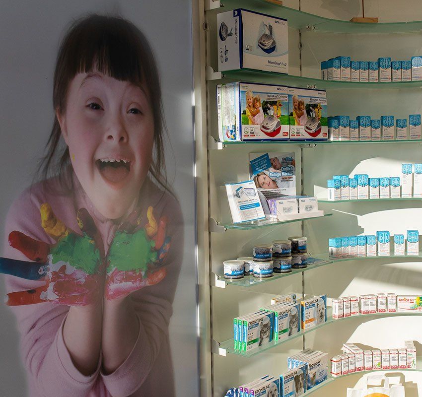 Ein Mädchen mit Farbe an den Händen lächelt strahlend neben einem Verkaufsregal voller Medikamentenpackungen.