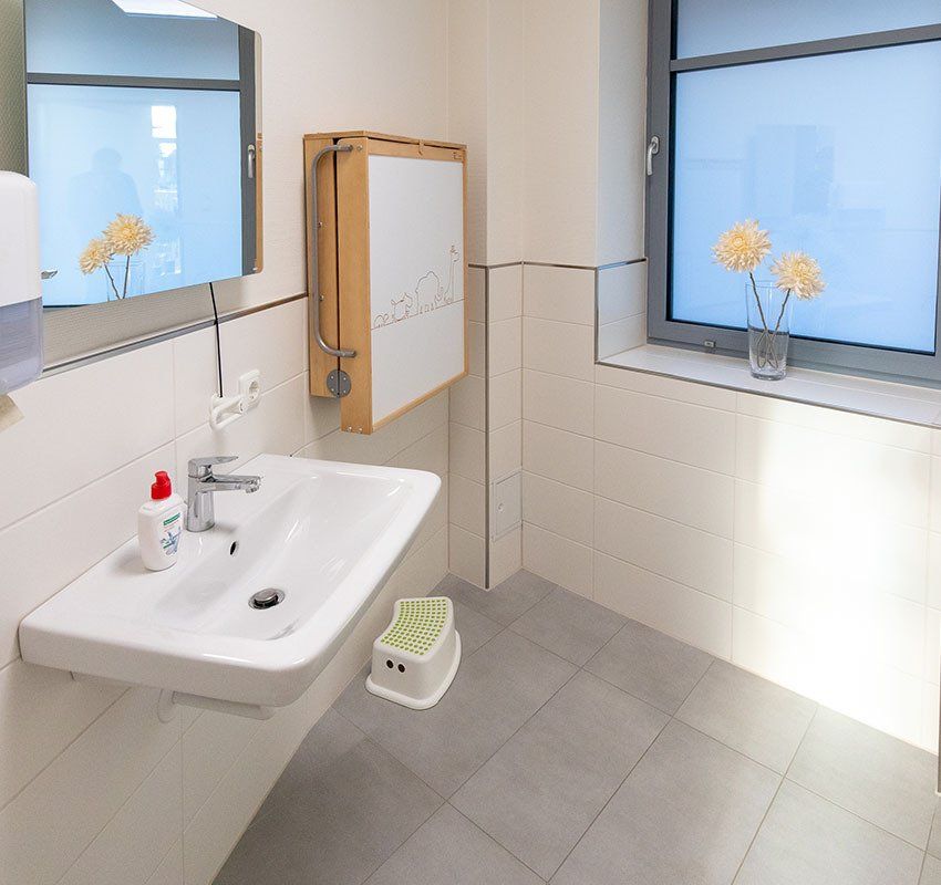 Ein modernes, barrierefreies Badezimmer mit weißem Waschbecken, einem an der Wand montierten, ausklappbaren Wickeltisch, einem kleinen Tritthocker und einem Fenster.