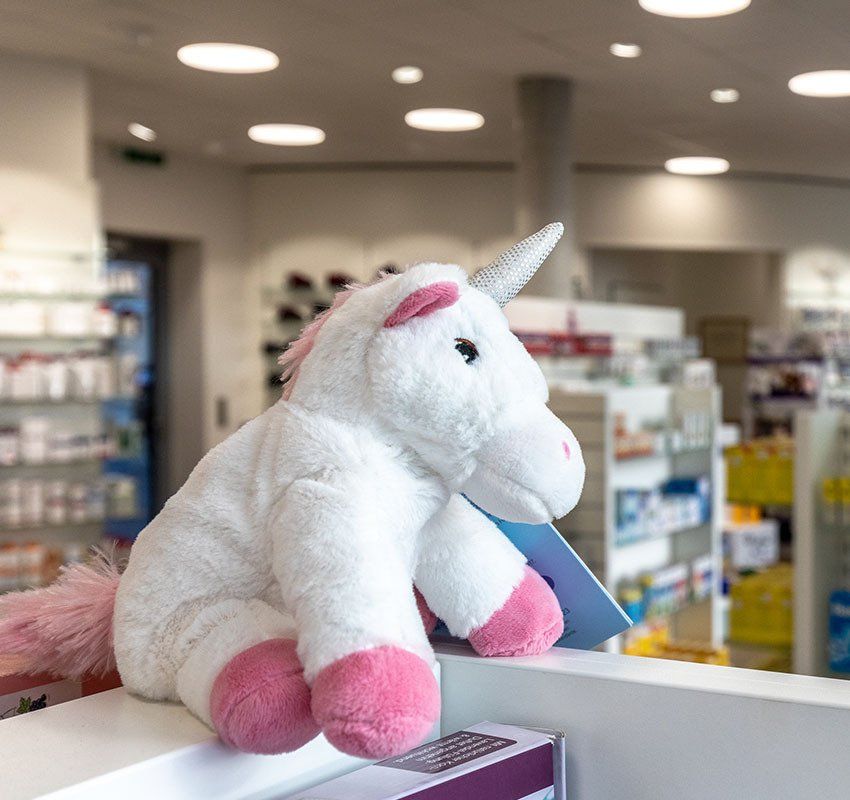 Ein weißes Plüscheinhorn mit rosa Hufen und rosa Mähne sitzt auf einem Tresen in einer Apotheke oder einem Geschäft.