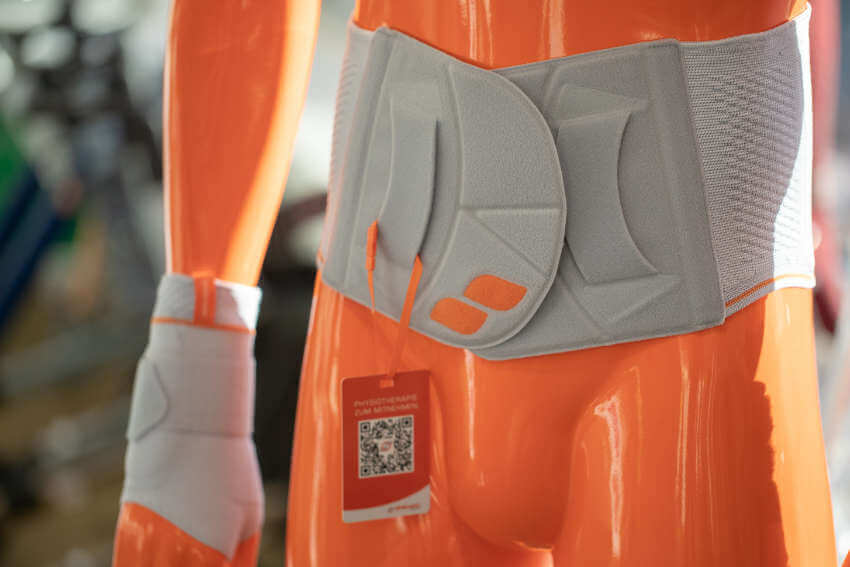 Eine orangefarbene Schaufensterpuppe trägt eine graue orthopädische Lendenwirbelstütze und eine dazu passende Handgelenkstütze mit einem angebrachten QR-Code-Etikett.