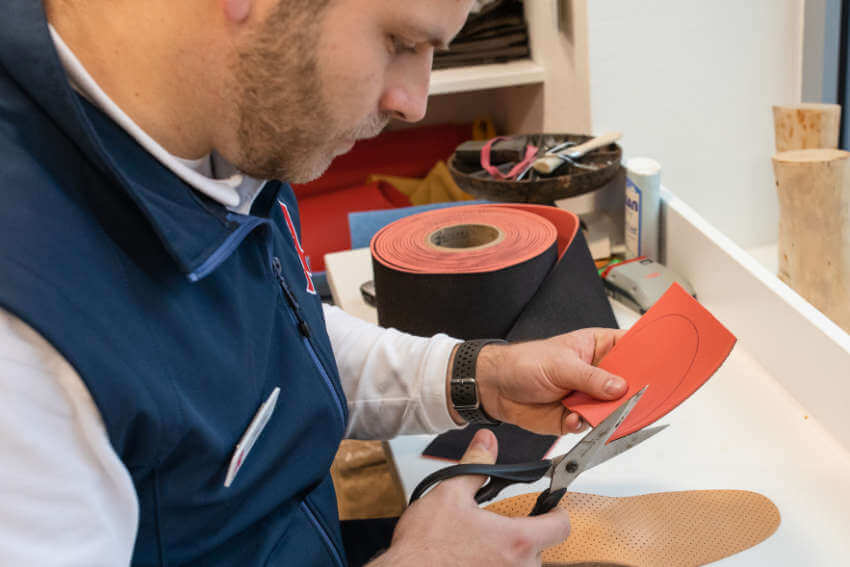 Eine Person in einer blauen Weste schneidet an einer Werkbank mit einer Schere einen roten Flicken aus; im Hintergrund ist eine Rolle roten Stoffs zu sehen.