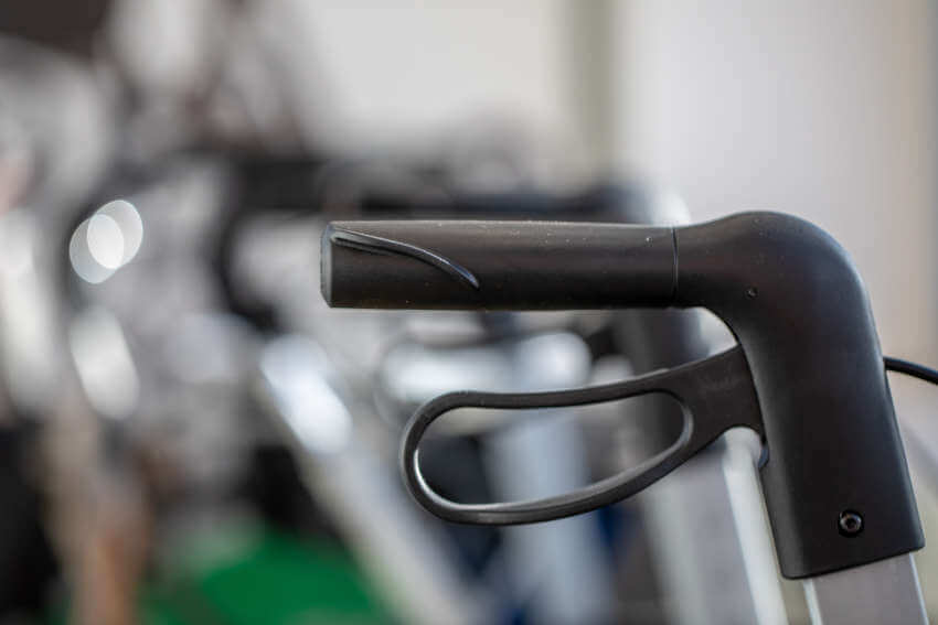 Nahaufnahme eines schwarzen, ergonomischen Handgriffs und eines Bremshebels in Schlaufenform an einem Rollator vor einem verschwommenen Innenraumhintergrund.