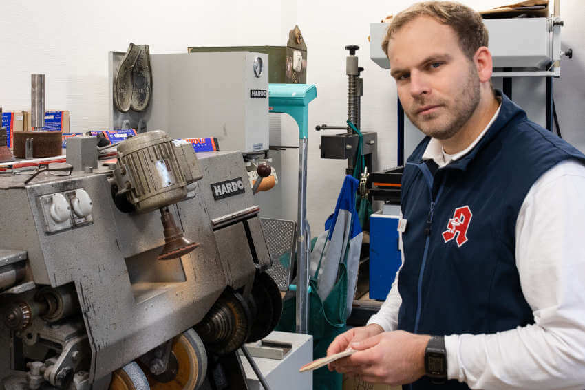 Eine Person in Apothekerweste hält in einer Werkstatt mit Industrieanlagen ein kleines Holzwerkzeug in der Hand.