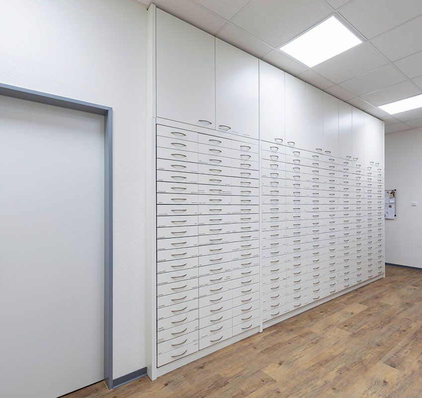 Eine weiße Wand mit Aufbewahrungsschränken und zahlreichen kleinen Schubladen in einem hellen Büroraum mit Holzbodenoptik.
