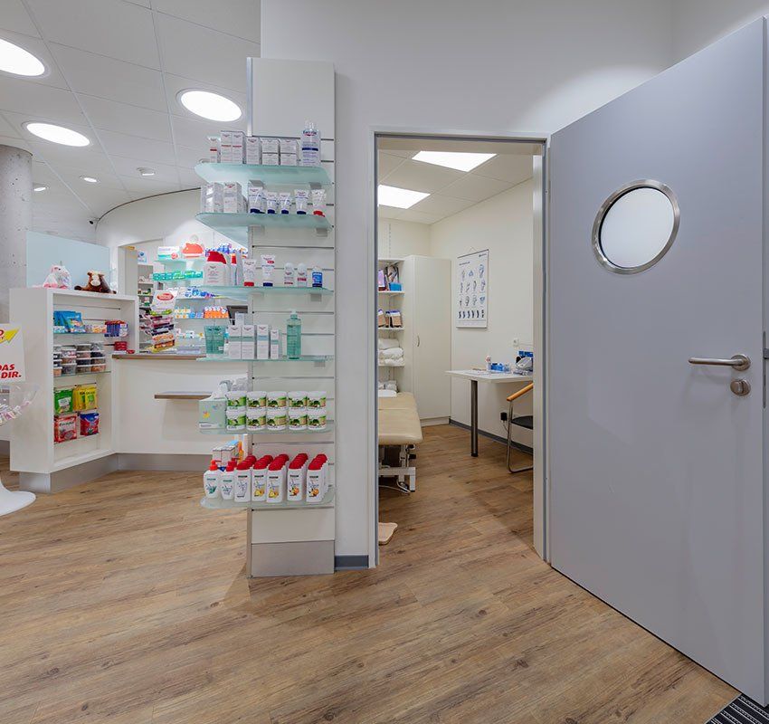 Ein lichtdurchfluteter Apothekeninnenraum mit Regalen voller Produkte, einem privaten Beratungsraum mit Bett und einer grauen Tür.
