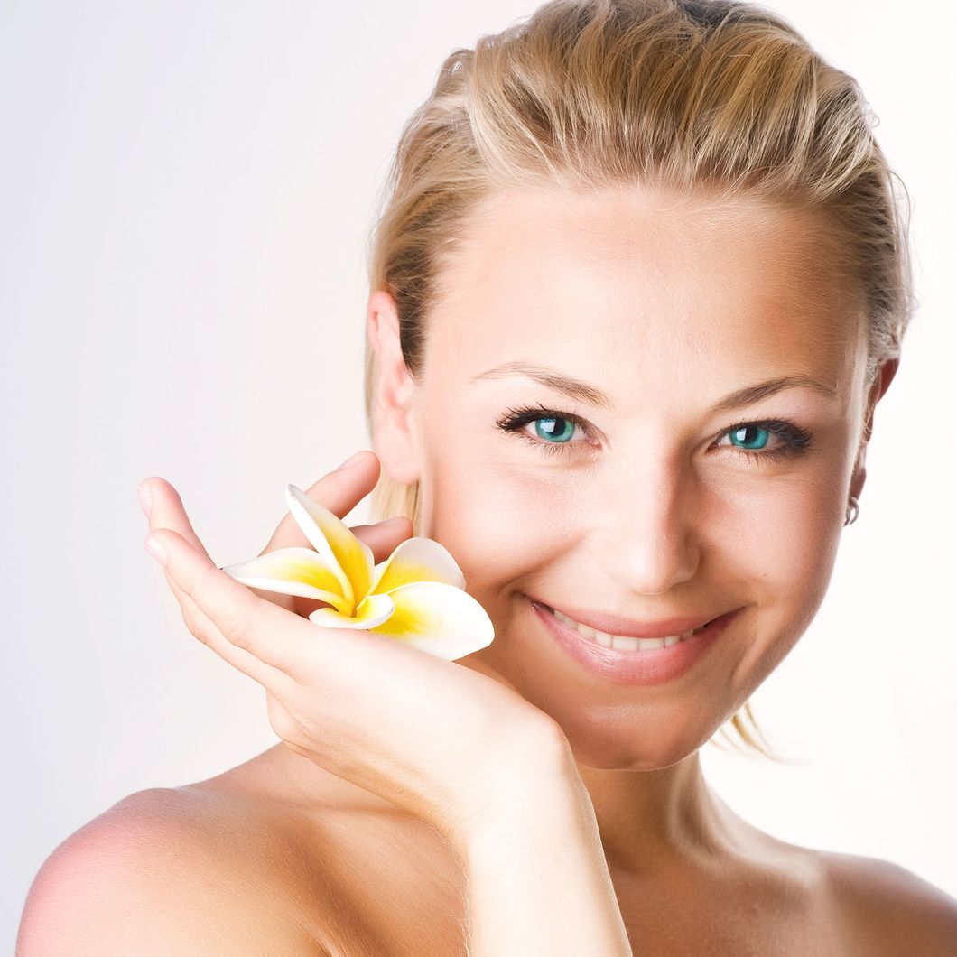 Eine lächelnde Person mit blonden Haaren hält vor einem weißen Hintergrund eine weiß-gelbe Frangipaniblüte vor ihr Gesicht.