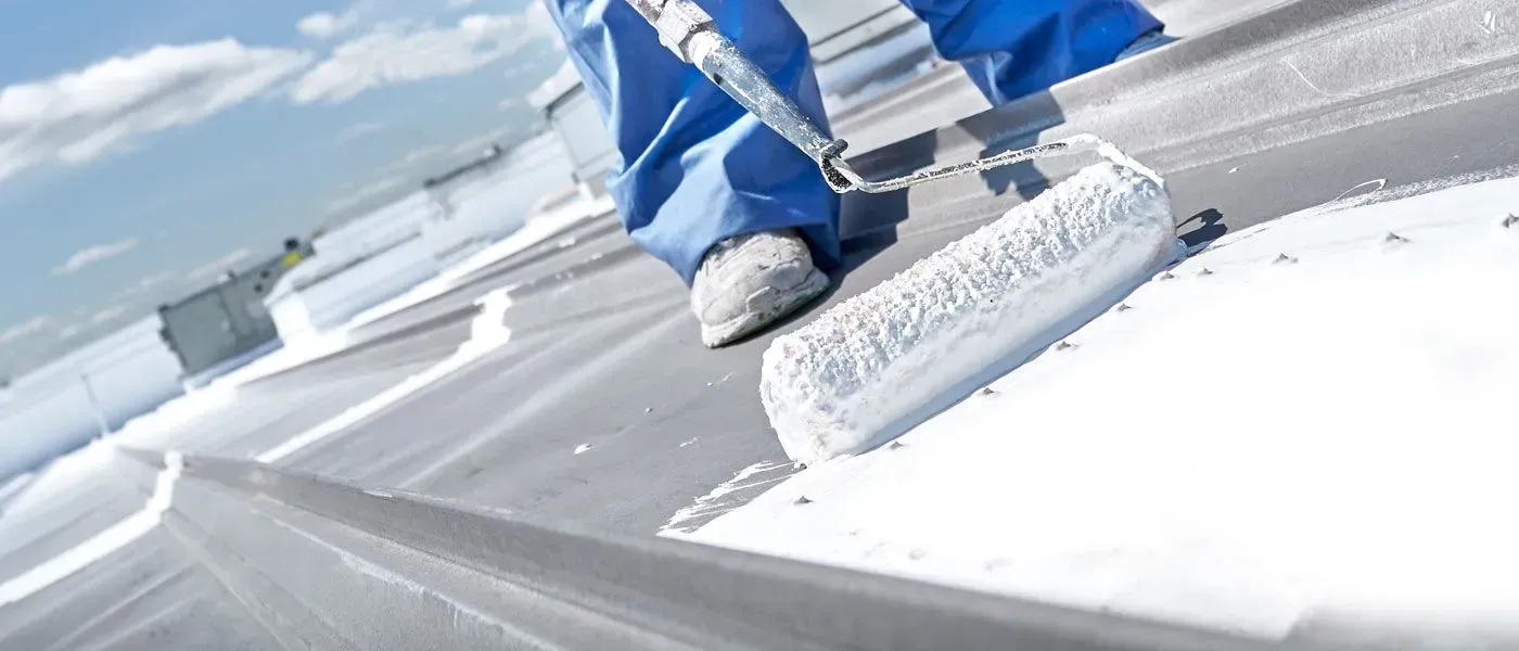 Person in blue overalls painting a gray metal roof with a white roller.
