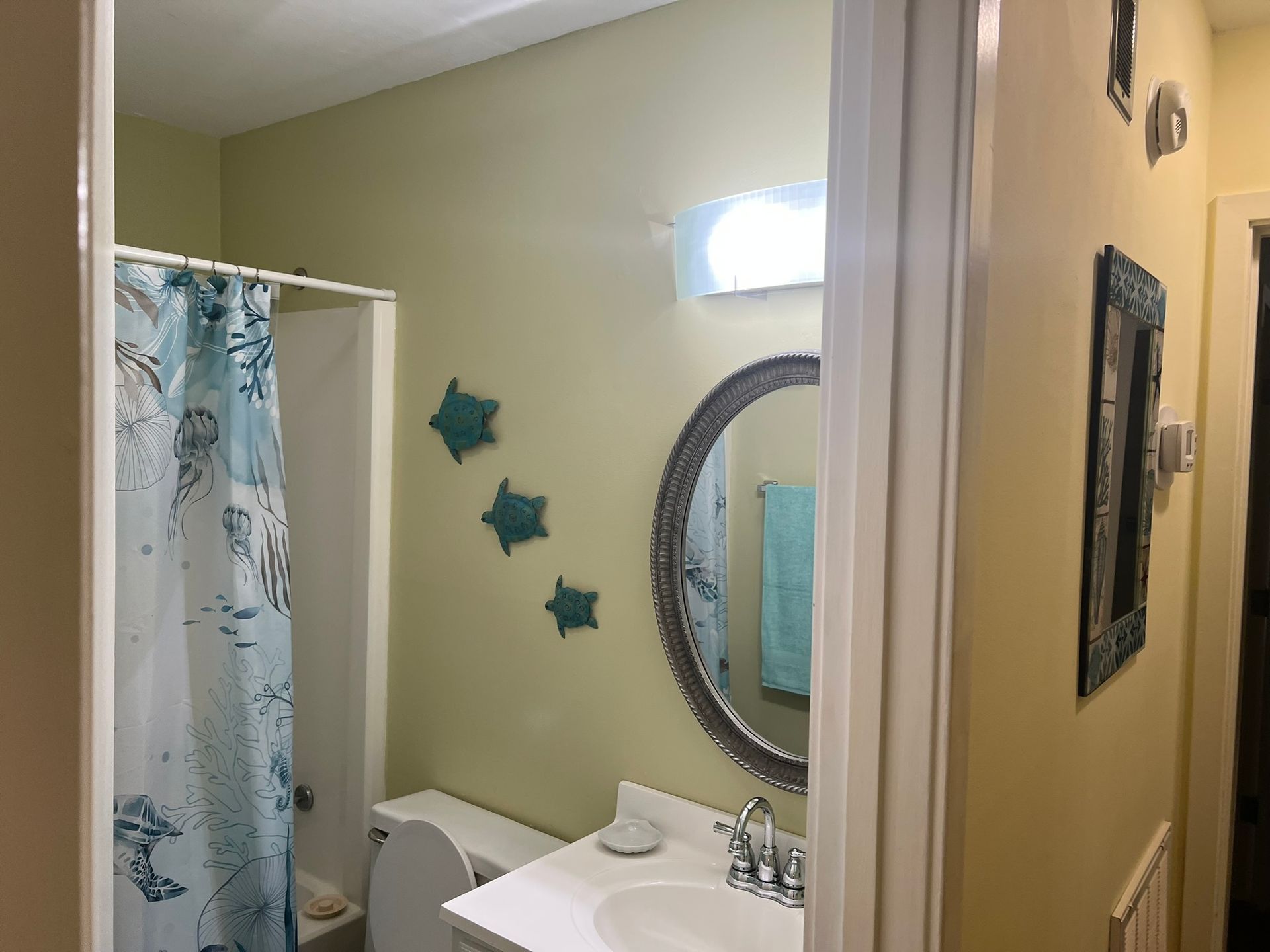 A bathroom with a white vanity, an oval mirror, teal turtle wall decor, and a blue and white patterned shower curtain.